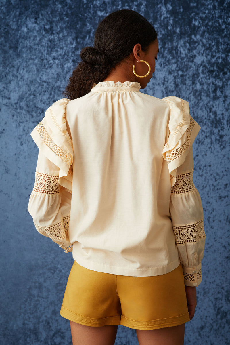 Blouse in a peach solid color with large puffed long sleeves with lace detailing wrapped around the sleeves and ruffles at the top of the shoulder